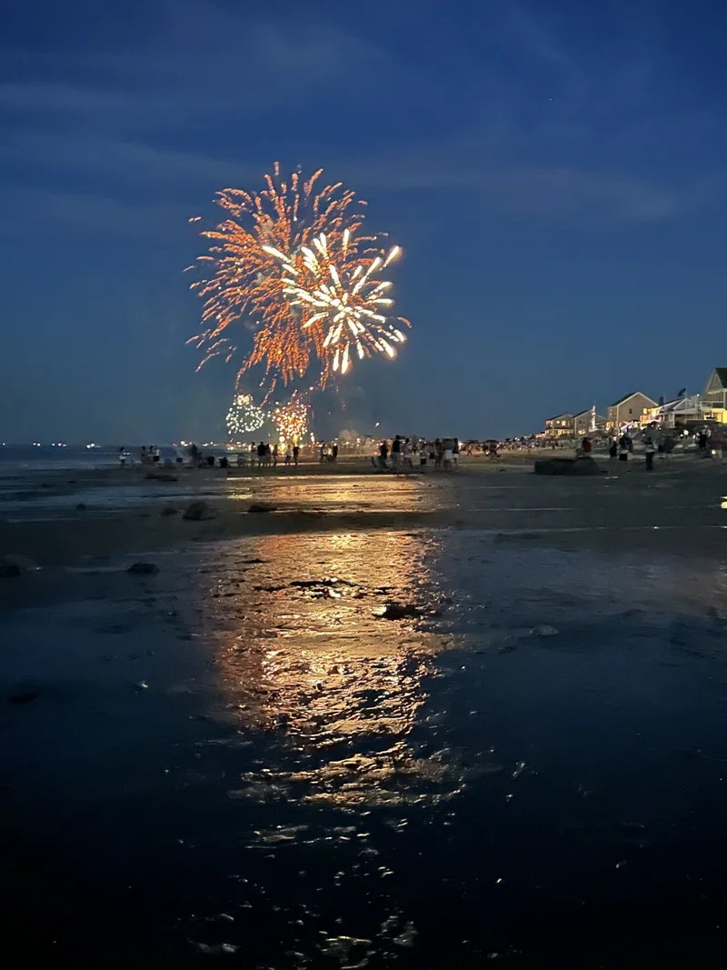 Fireworks over a South Shore beach on the night of July 3rd, reflected on the wet sand as neighbors watch from the shoreline